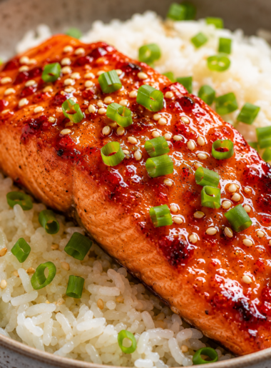 A close-up shot of a baked Thai sweet chili salmon fillet served over a bed of white jasmine rice, garnished with green onions and toasted sesame seeds.