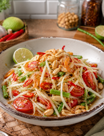 Thai Green Papaya Salad (Som Tum) served in a ceramic bowl on a kitchen counter, featuring shredded green papaya, cherry tomatoes, green beans, roasted peanuts, dried shrimp, and red chilies, with fresh ingredients and a rustic kitchen background.
