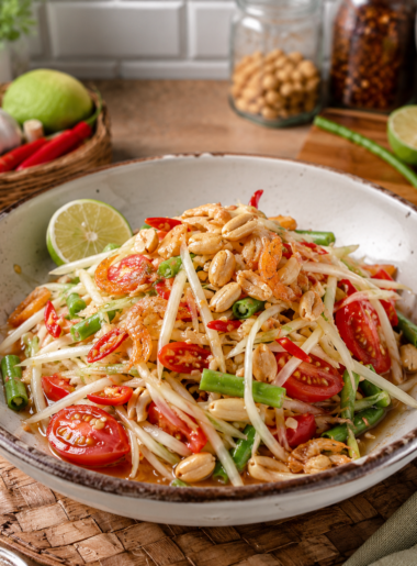 Thai Green Papaya Salad (Som Tum) served in a ceramic bowl on a kitchen counter, featuring shredded green papaya, cherry tomatoes, green beans, roasted peanuts, dried shrimp, and red chilies, with fresh ingredients and a rustic kitchen background.
