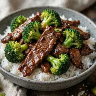 Chinese beef and broccoli stir-fry served over steamed rice in a bowl, with glossy sauce, tender beef strips, and bright green broccoli florets.